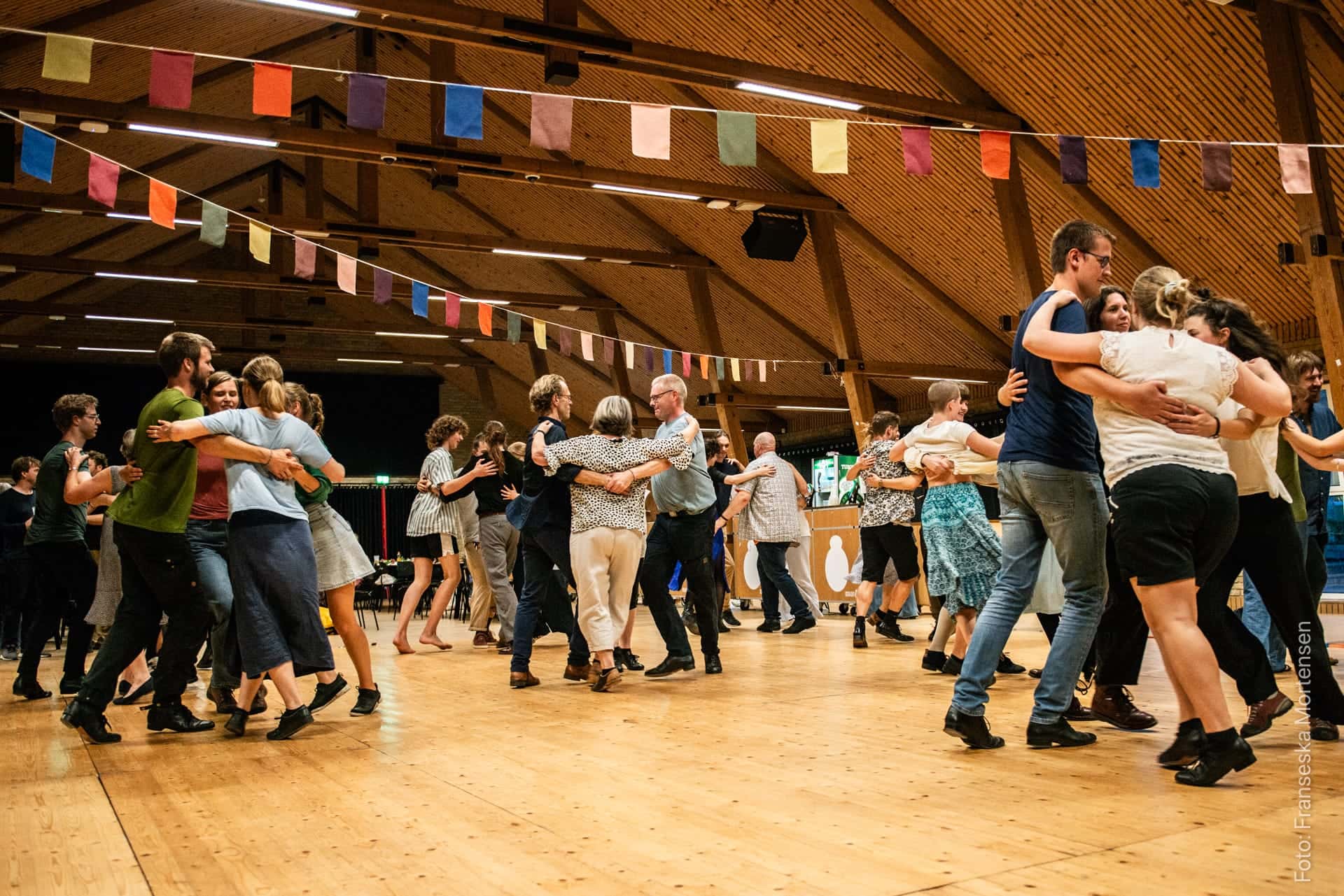 Festivalgæster danser sammen under farverige bannere i en stor træsal