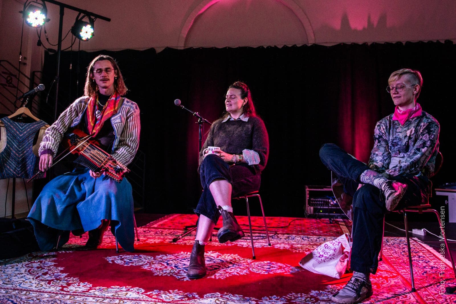 Fredy Clue with a nyckelharpa and two conversation partners seated on a red rug on the FOLK stage