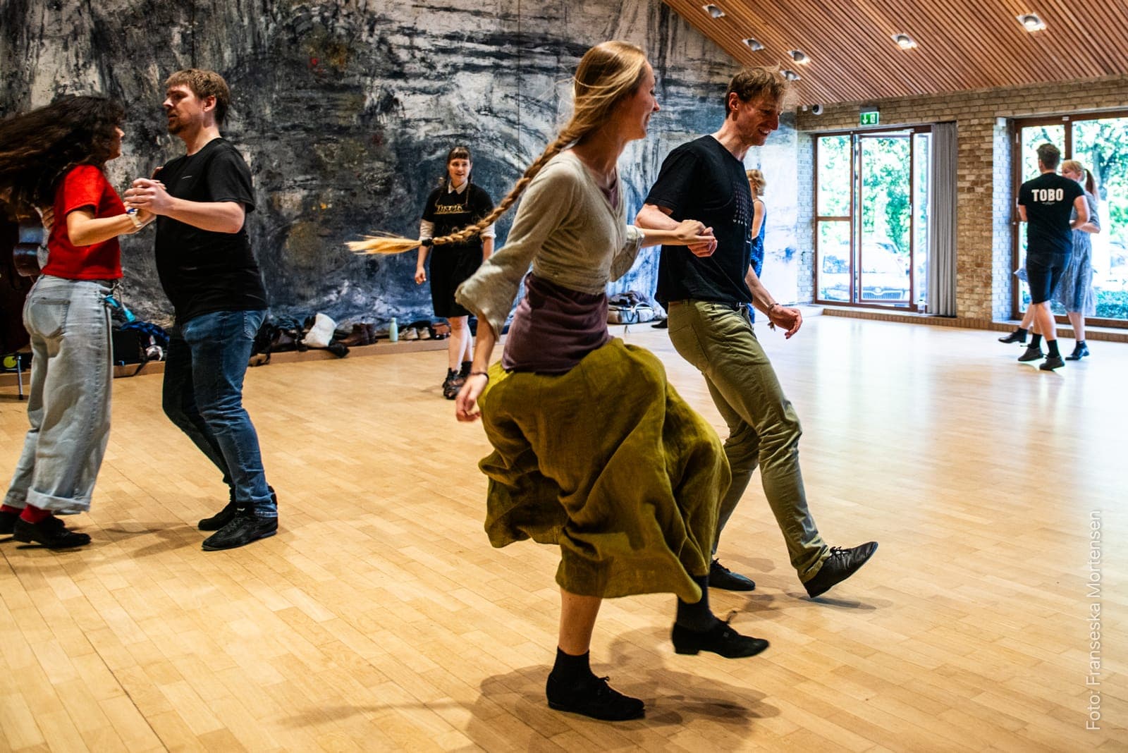 Couples dancing polka across the wooden floor, with large windows and a mural behind them