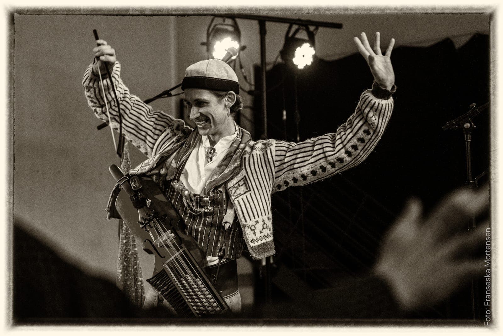 Black-and-white photo of Fredy Clue in a striped jacket with a nyckelharpa, laughing with arms raised