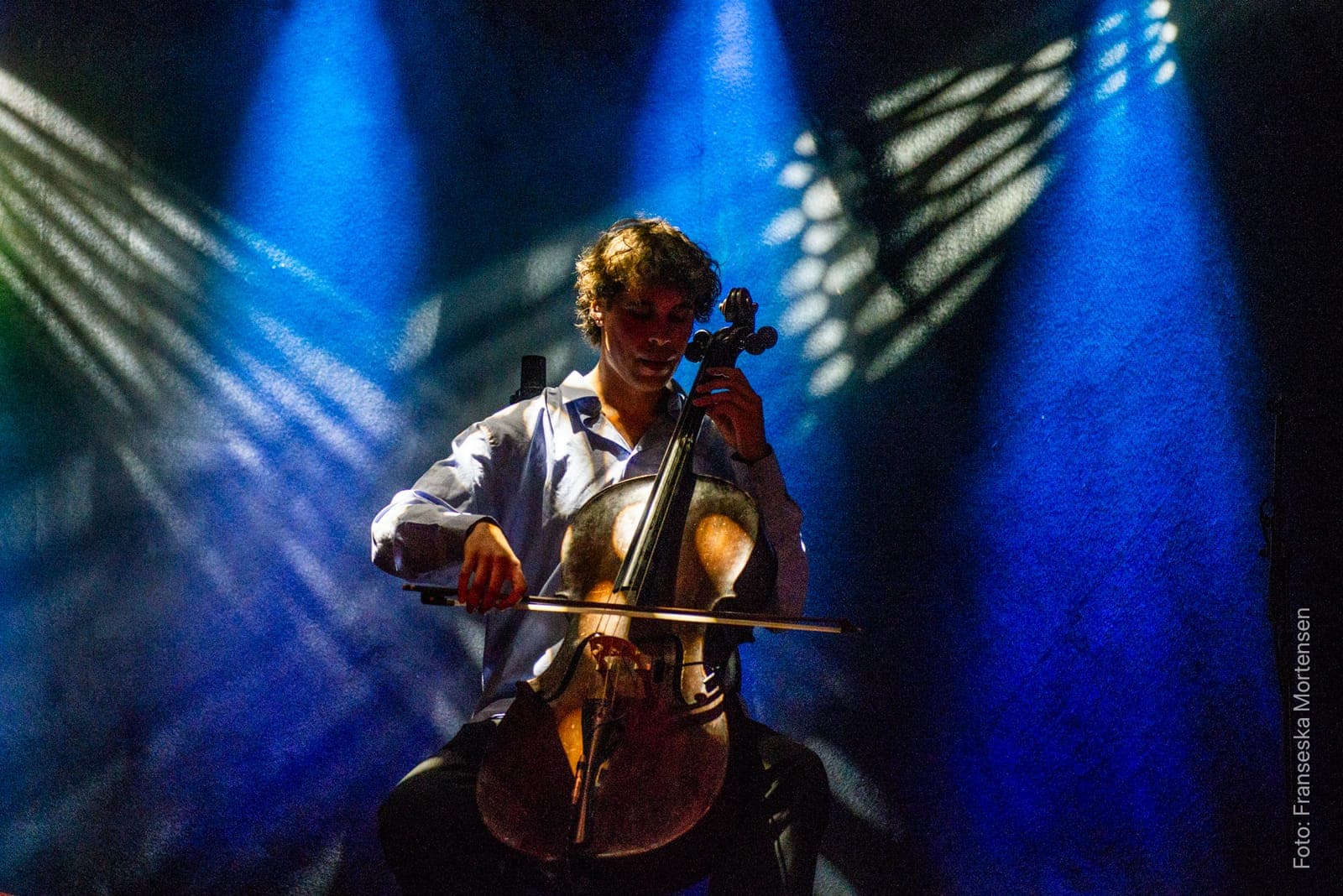 Asbjørn Elle of Geo Pjäserna playing cello in blue light, with patterned beams of light behind