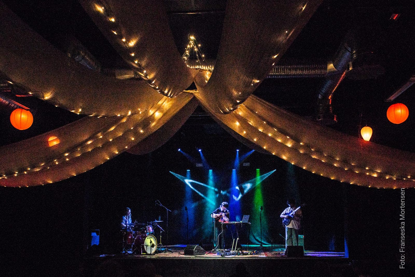 Geo Pjäserna playing as a trio on the turkis stage beneath white fabric drapes with string lights and red lanterns