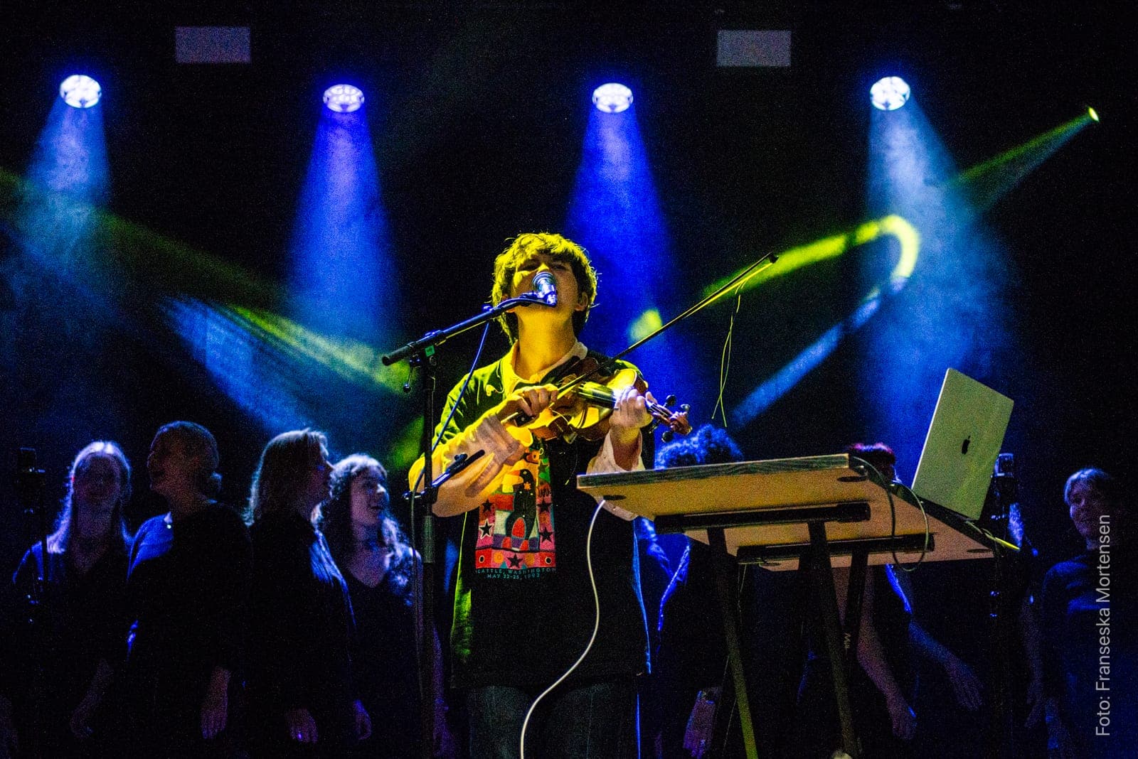 Julia Bengtson singing and playing the violin in front of a choir in blue and yellow stage light