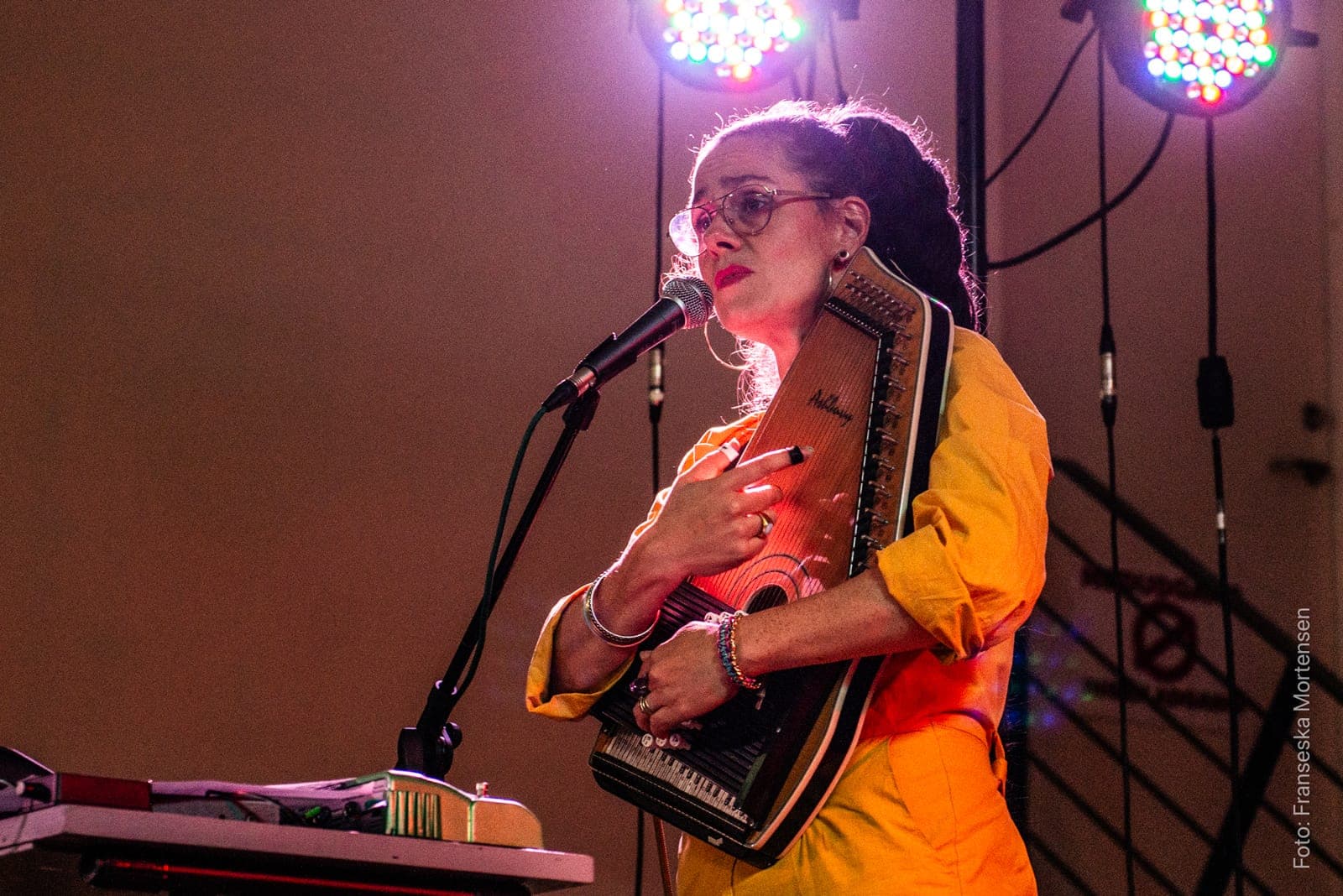Jullie Hjetland in a yellow jumpsuit singing into the microphone, holding an autoharp