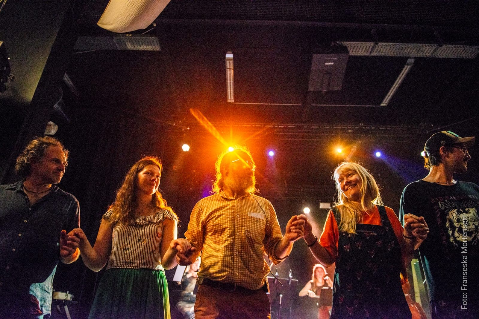 Dancers holding hands in a chain and smiling in orange and blue stage light