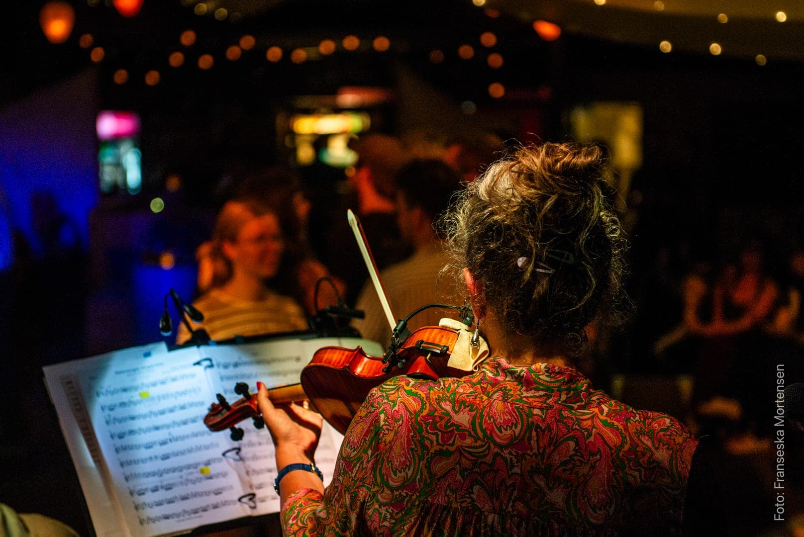 A violinist from Det Lyse Bal seen from behind with the sheet music, and dancers blurred in the background
