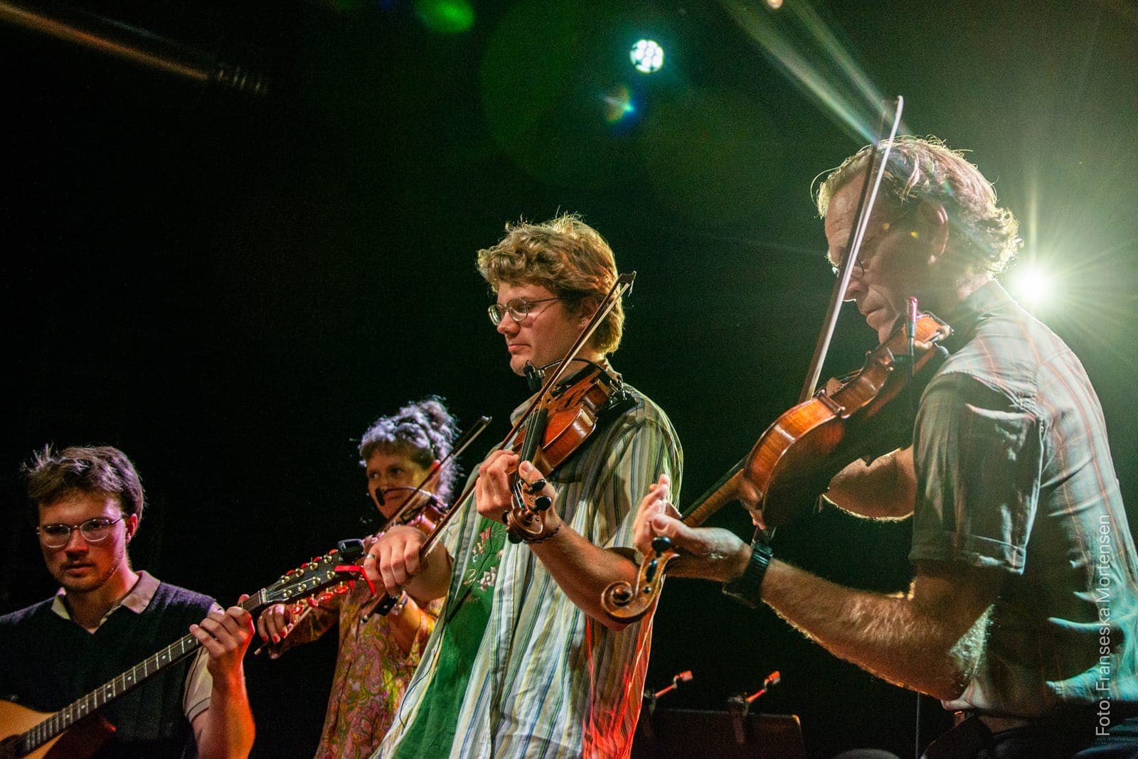 Three fiddlers and a guitarist playing close together on stage in green backlight