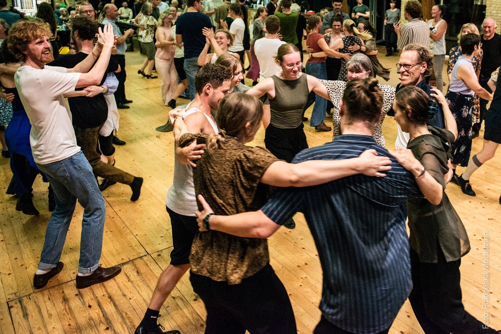 Dansere i alle aldre danser i små kredse i en fyldt sal ved en tidligere festival