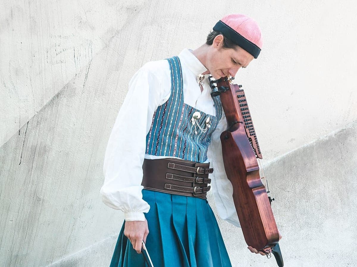 Fredy Clue står med nyckelharpa og bue iført den blå Bäckadräkten