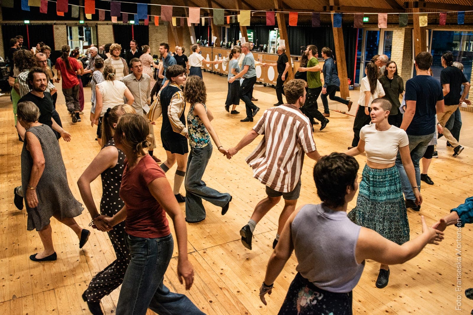 Fyldt dansegulv ved et tidligere KOM FOLK DANS, hvor dansere i alle aldre holder hinanden i hænderne i kæder
