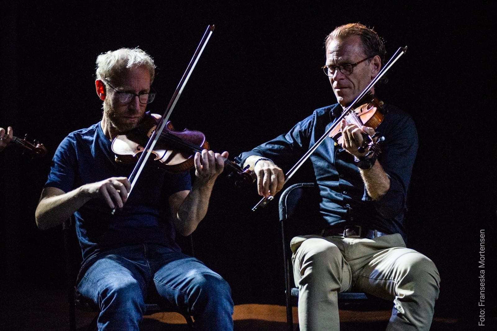 To violinister spiller tæt sammen på en mørk scene.